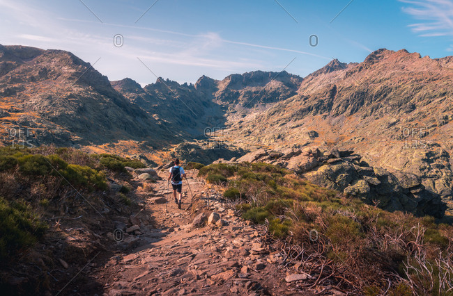 From above back view of unrecognizable male tourist with rucksack trekking with poles on rough pathway in mountains under sky
