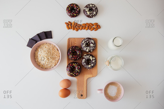 Top view composition with sweet donuts with chocolate topping and sprinkles placed on wooden board on white table with ingredients for recipe