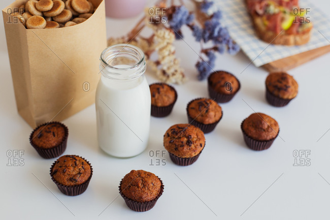 High angle composition with tasty baked sweet muffins with chocolate drops in paper cups arranged around glass bottle of milk on table with various snacks for breakfast