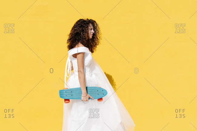 Side view of bride in white dress standing while holding longboard against yellow background in sunlight