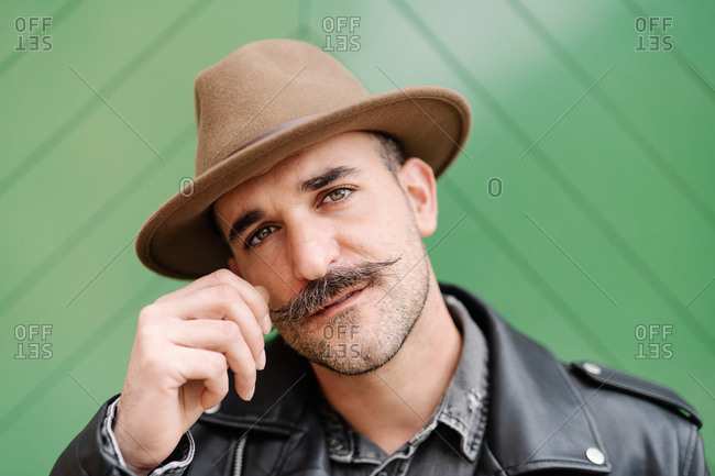 Mature male in leather jacket curling mustache while looking at camera standing on green gate