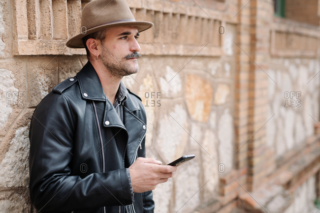 Side view of bearded male with mustache in hat and leather jacket using mobile phone and sending voice message on blurred background of building looking away