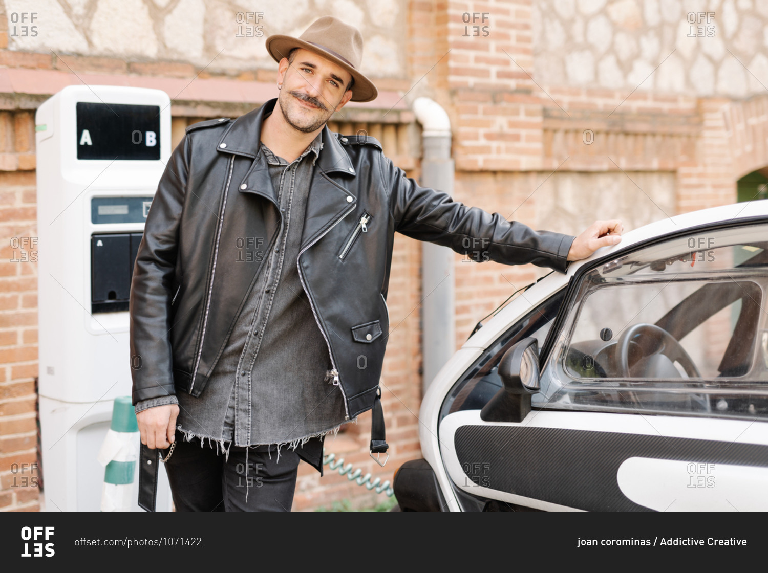 Trendy happy brutal male in hat standing near contemporary electric automobile on charge station near brick stone wall