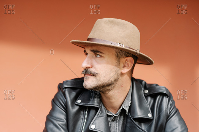 Trendy thoughtful male in hat and leather jacket on beige background looking away