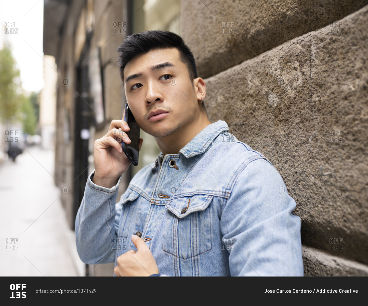 Serious young Asian male in casual outfit speaking on mobile phone while standing near stone wall of urban building