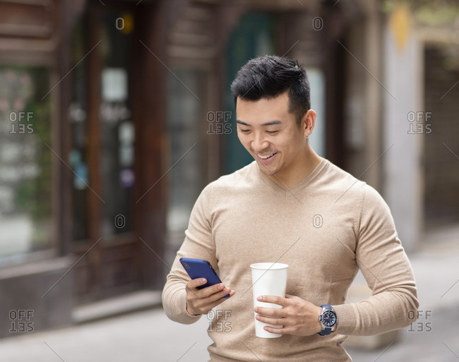 Smiling Asian male standing on street with hot aromatic drink to go and reading messages on social media via mobile phone