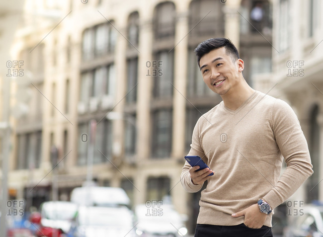 Positive masculine Asian male standing with hand in pocket and browsing mobile phone in city while looking away
