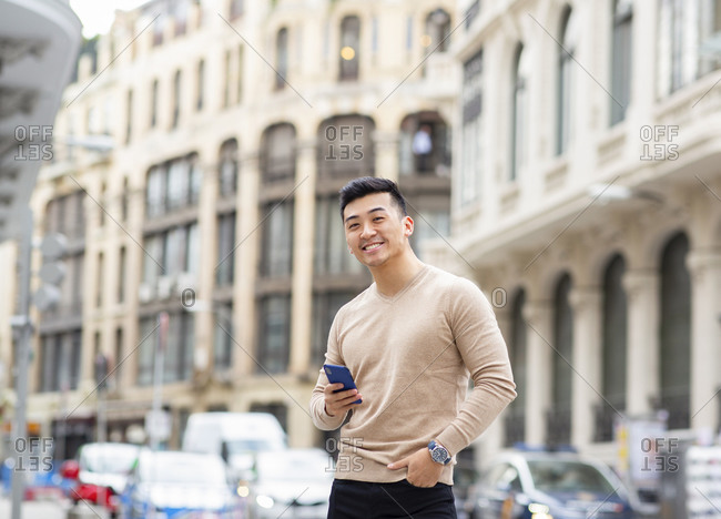 Positive masculine Asian male standing with hand in pocket and browsing mobile phone in city while looking at camera