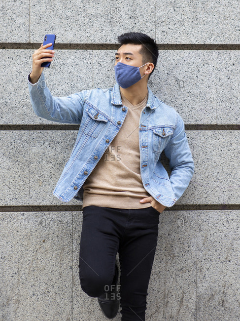 Asian male wearing medical protective mask standing near building and taking selfie on smartphone during coronavirus epidemic in city