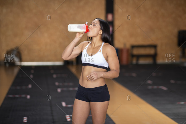 Young fit female athlete in active wear drinking sports beverage from plastic bottle after training in gymnasium and looking up