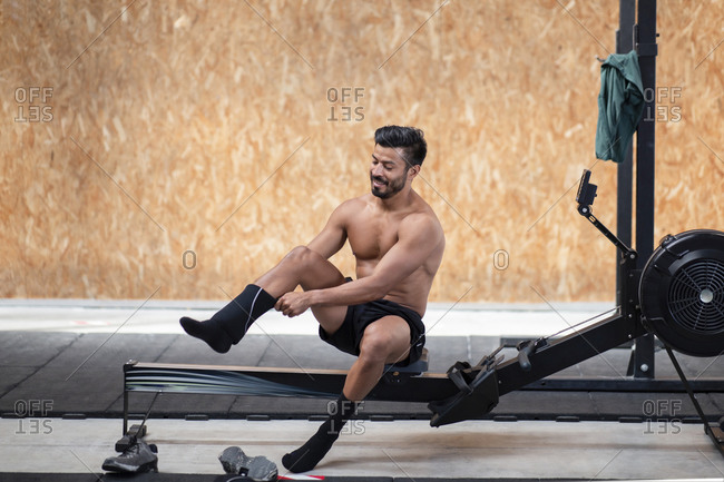 Cheerful unshaven muscular ethnic sportsman with naked torso putting on knee pad while sitting on bench before working out in gym and looking down