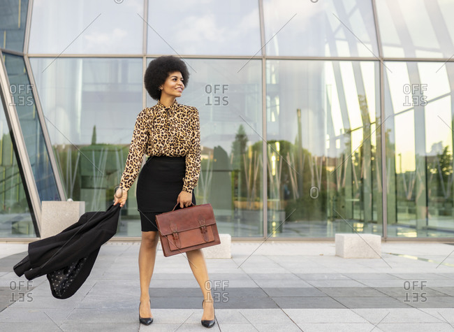 Full body of cheerful stylish African American female employee with afro hair and briefcase holding jacket in hands standing near glass walled building