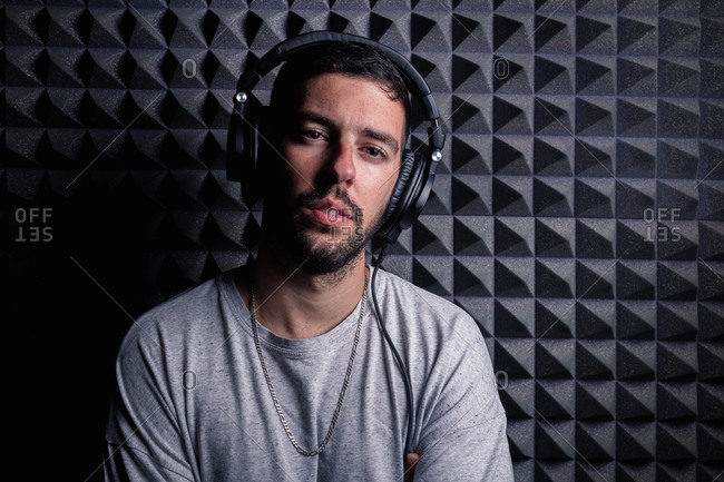 Stylish male singer wearing headphones standing in recording studio on background of soundproof acoustic panel and looking at camera
