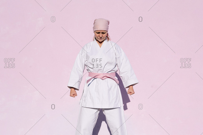Determined thoughtful mature woman in pink head cover and belt fighting karate in cancer battle concept in the street on pink wall with eyes closed