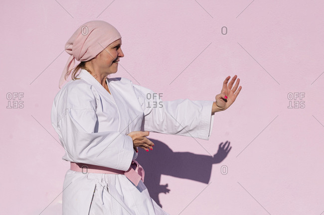 Side view of determined mature woman in pink head cover and belt fighting karate in cancer battle concept in the street on pink wall looking away