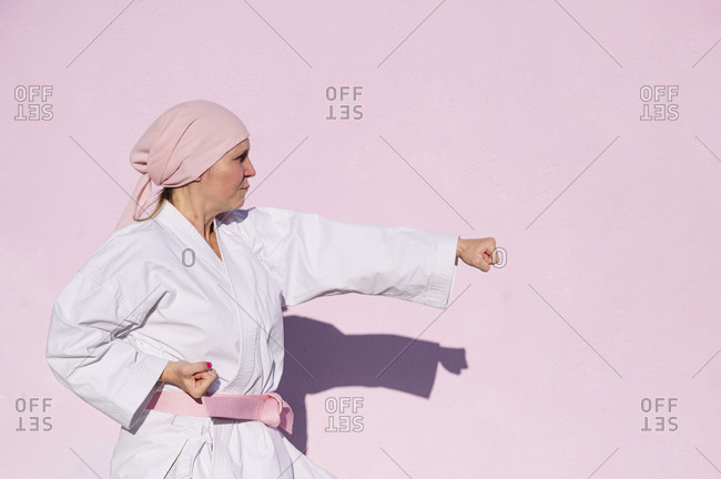 Side view of determined mature woman in pink head cover and belt fighting karate in cancer battle concept in the street on pink wall looking away