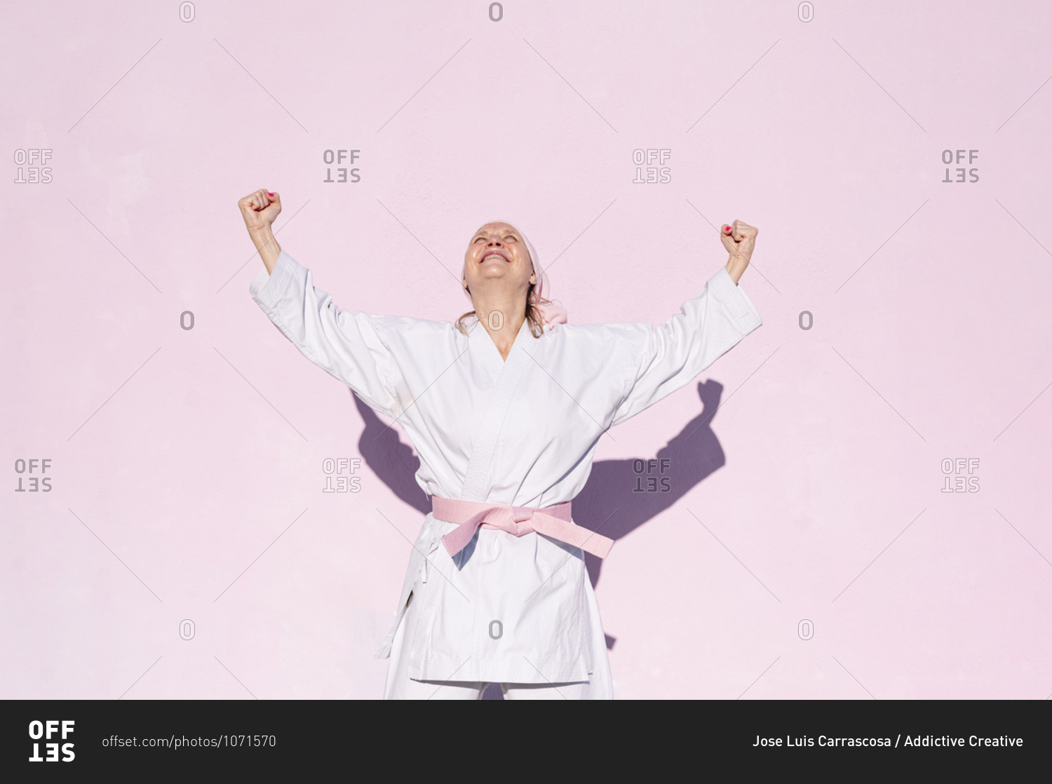 Determined mature woman in pink head cover and belt fighting karate in cancer battle concept doing victory gesture in the street on pink wall looking up