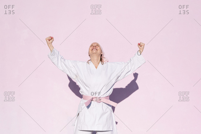 Determined mature woman in pink head cover and belt fighting karate in cancer battle concept doing victory gesture in the street on pink wall looking up