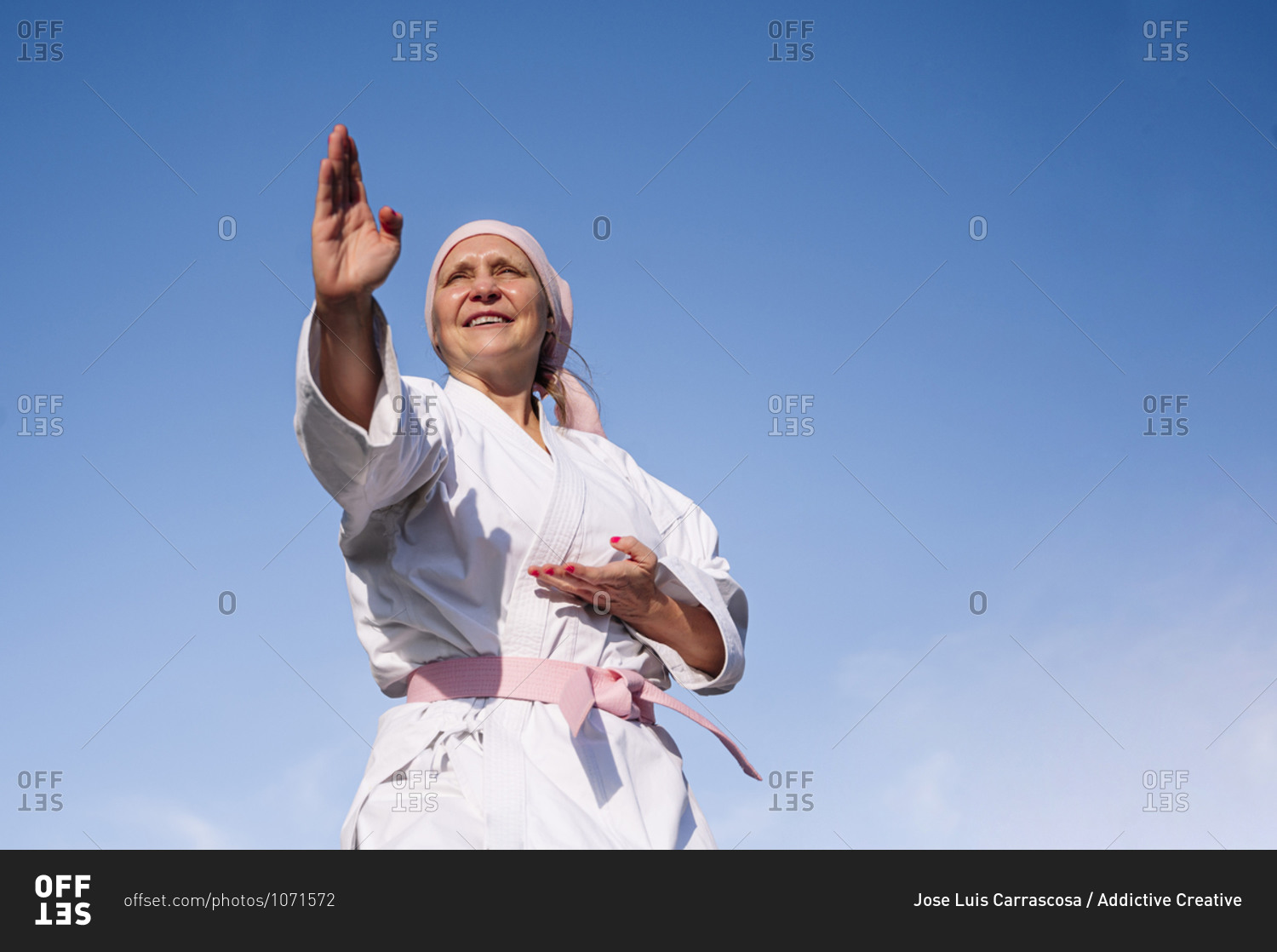 From below determined mature woman in pink head cover and\
belt fighting karate in cancer battle concept standing on blue sky\
background looking away stock photo - OFFSET