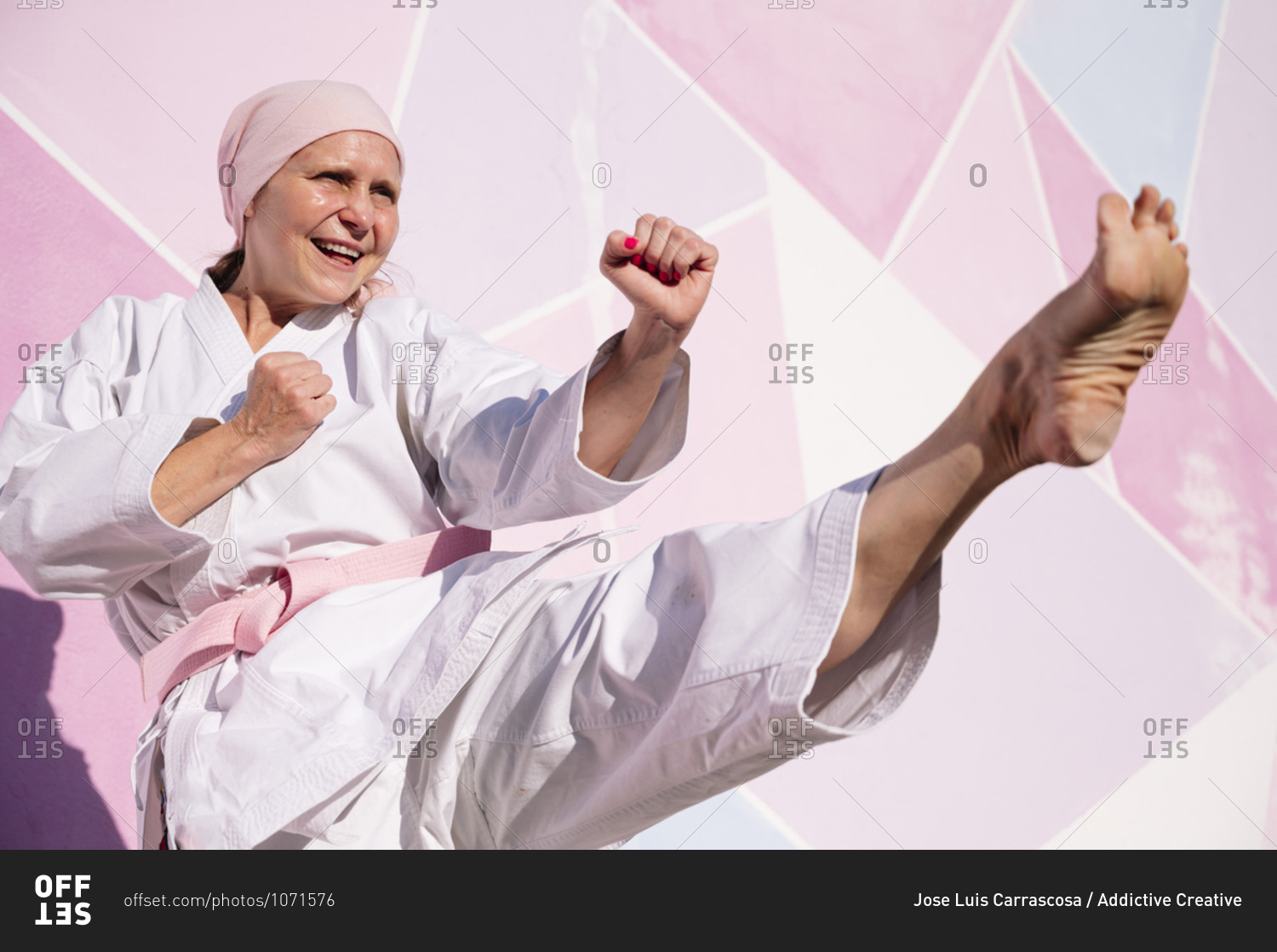 Determined cheerful mature barefooted woman in pink head cover and belt kicking fighting karate in cancer battle concept in the street on pink wall looking away