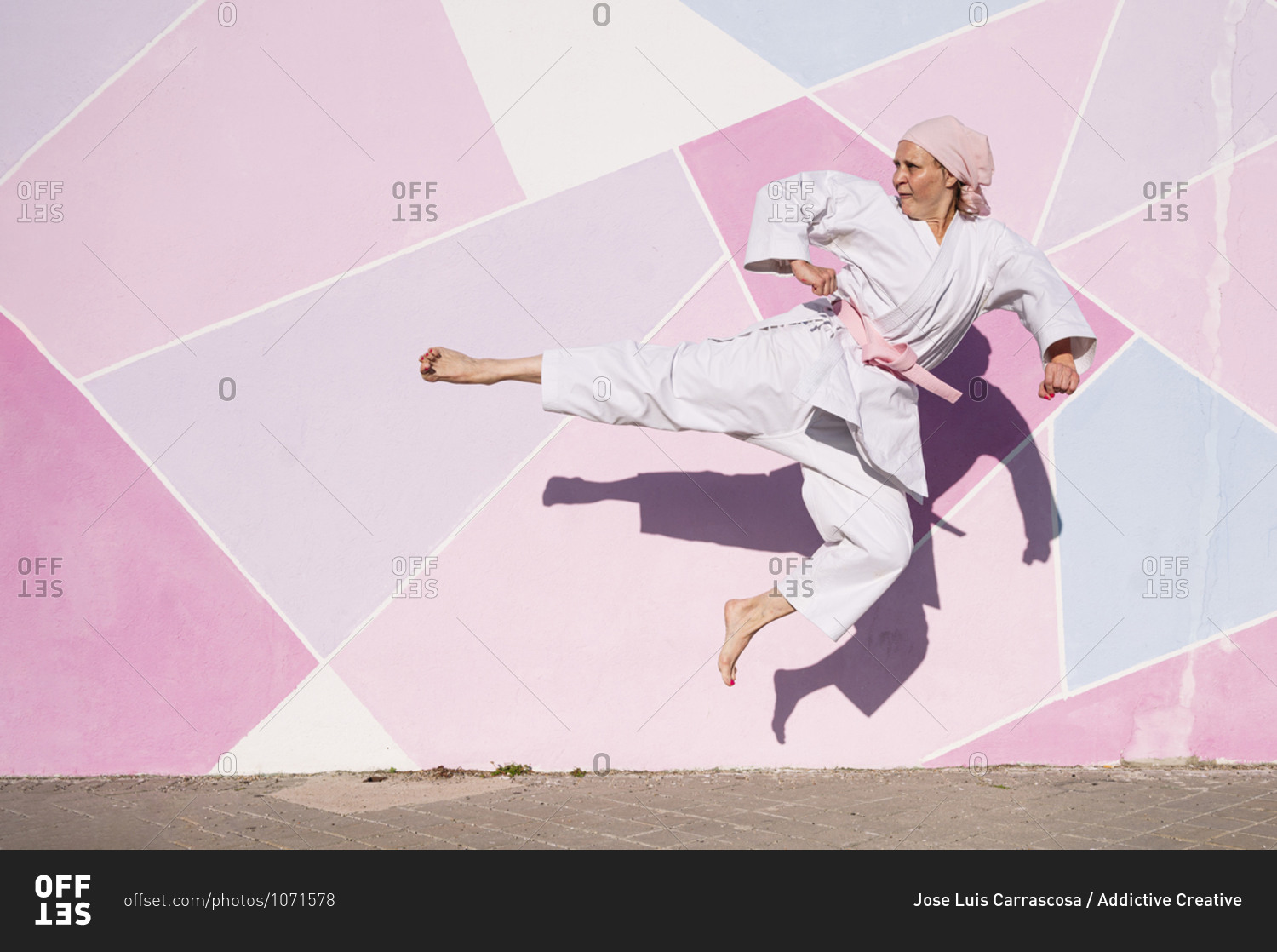 Determined mature barefooted woman in pink head cover and belt kicking fighting karate in cancer battle concept in the street on pink wall looking away