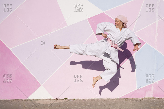 Determined mature barefooted woman in pink head cover and belt kicking fighting karate in cancer battle concept in the street on pink wall looking away