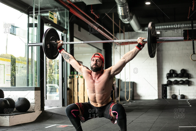 Muscular focused shirtless male athlete doing clean and jerk exercise with barbell during weightlifting training in gym looking away