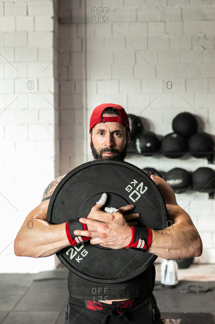 Muscular sportsman in weight vest and with heavy disc doing squats during workout in gym looking at camera