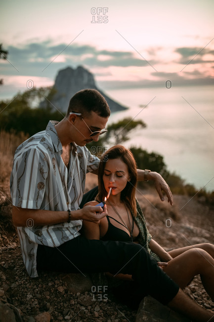 Side view of calm man lighting cigarette for girlfriend while chilling on seashore at sunset