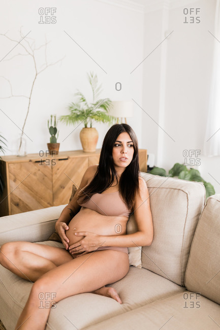 Female expecting baby and wearing lingerie sitting in cozy couch and touching tummy looking away