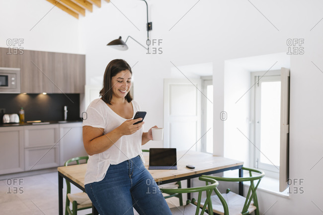 Smiley woman typing in a smartphone with a coffee in a modern apartment