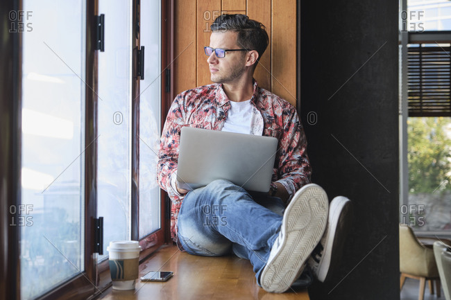Young trendy remote worker in eyeglasses with netbook sitting with crossed legs on windowsill near disposable glass of coffee while looking out window