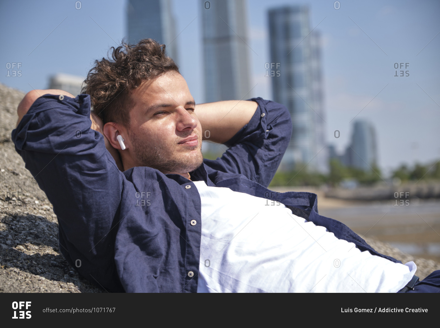 Content stylish young male with hands behind head listening to music in modern earphones while lying on stone near city river and looking away
