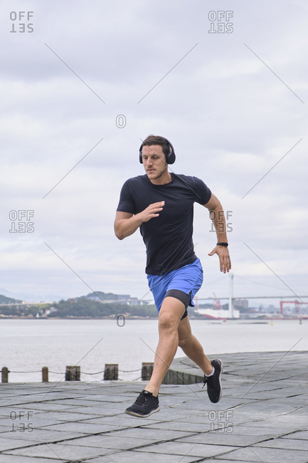 Ground level of focused male runner warming up and jumping above ground during training in city on cloudy day