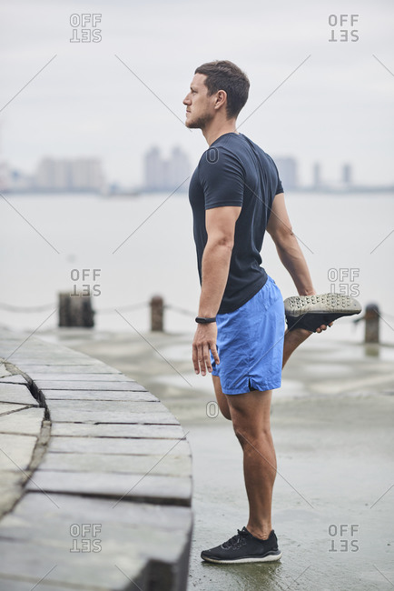 Concentrated male runner in sportswear standing on embankment and warming up while stretching legs and doing forward bend during workout