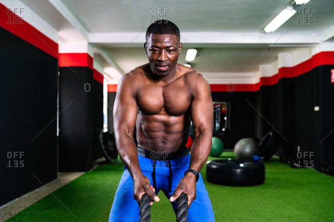 Ethnic sportsman with strong torso exercising with battle ropes during functional training in gym
