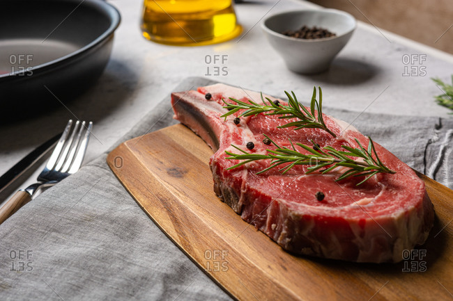 Uncooked t bone beef steak garnished with black pepper and rosemary sprigs placed on wooden chopping board in kitchen
