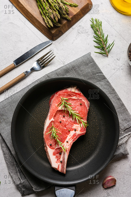 Top view of uncooked raw t bone steak garnished with rosemary and black pepper peas placed in frying pan on table in kitchen