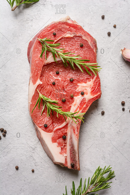 Top view uncooked t bone beef steak garnished with black pepper and rosemary sprigs placed on table in kitchen