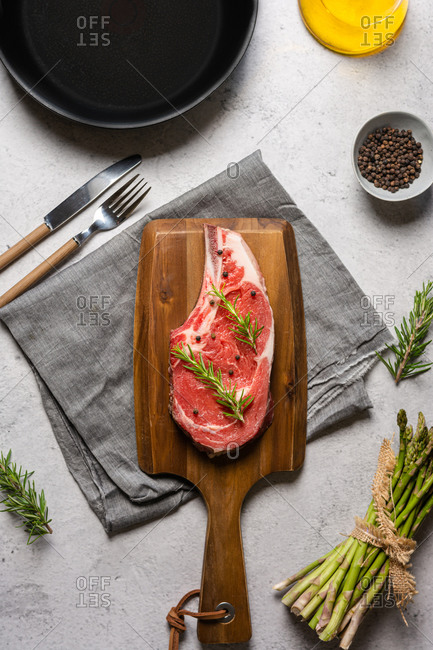 Top view uncooked t bone beef steak garnished with black pepper and rosemary sprigs placed on wooden chopping board in kitchen