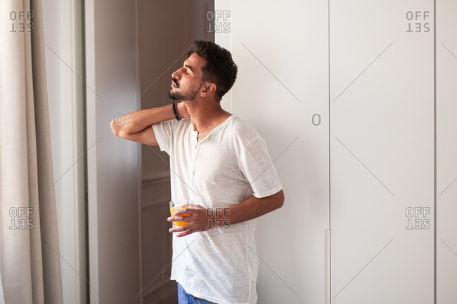 Side view of young male in casual clothes drinking fresh juice and observing street while standing near window in modern apartment in morning with eyes closed