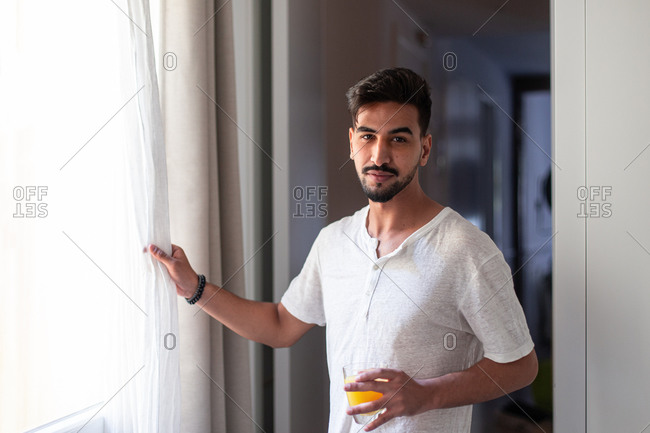 Side view of young male in casual clothes drinking fresh juice and observing street while standing near window in modern apartment in morning looking at camera