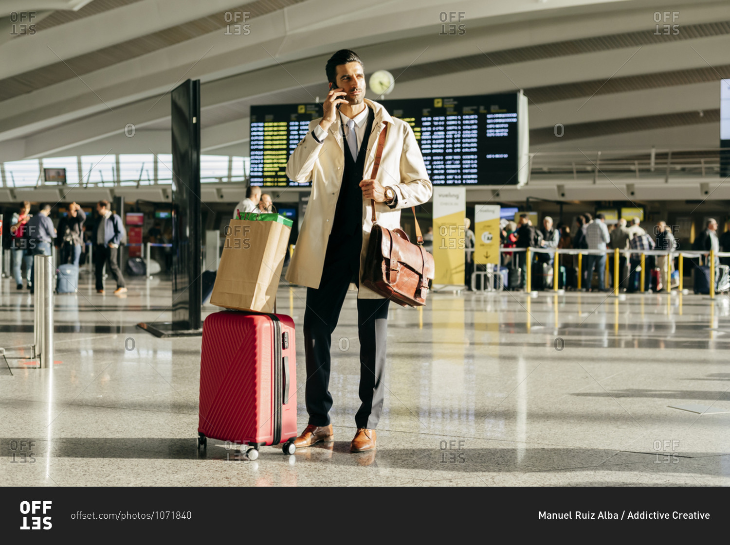 Man with dark hair in stylish clothes talking on phone while standing with suitcase in terminal of airport
