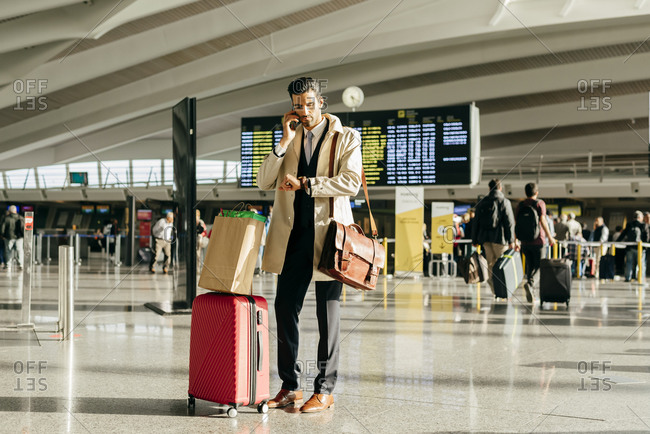 Man with dark hair in stylish clothes talking on phone and looking at watch while standing with suitcase in terminal of airport