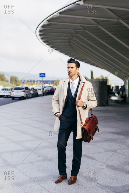 Man with dark hair in suit and coat looking away while standing near airport terminal at daytime