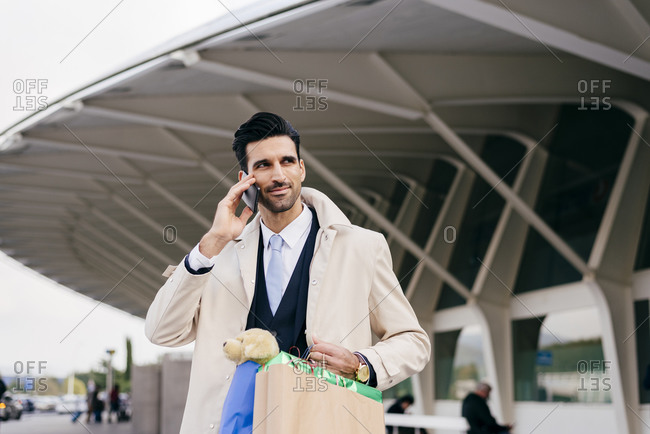 Positive man with black hair in formal clothes standing with shopping bags and talking on phone against modern building