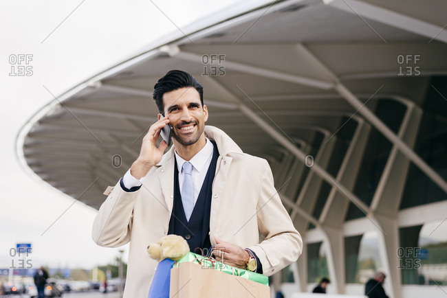 Positive man with black hair in formal clothes standing with shopping bags and talking on phone against modern building