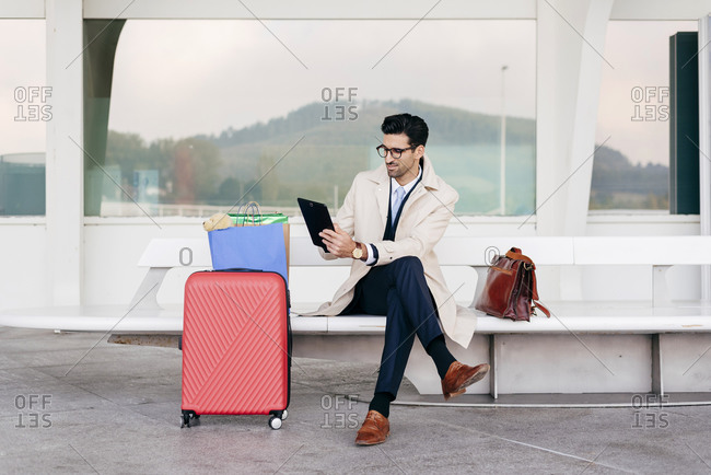 Confident man with hairstyle in stylish formal clothes with suitcase and shopping bags sitting on bench near airport and using tablet