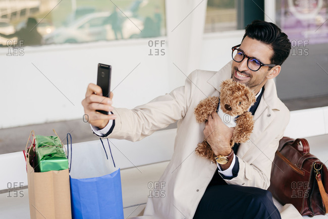 Smiling man with hairstyle in trendy formal clothes sitting on bench with suitcase and shopping bags and holding toy dog and taking selfie against modern building at daytime