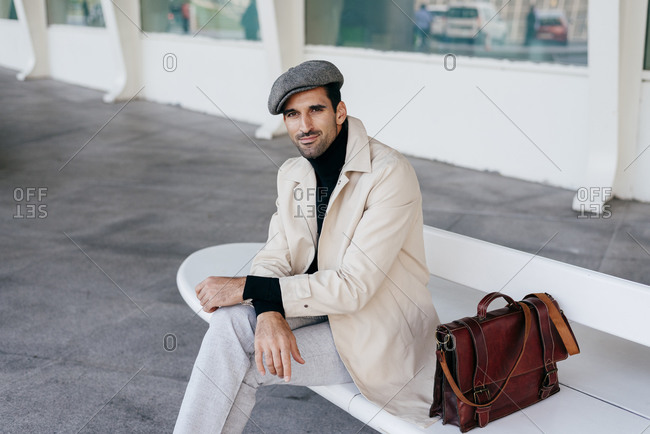Confident man with beard in trendy clothes sitting on white bench with bag and looking at camera against modern building with big window